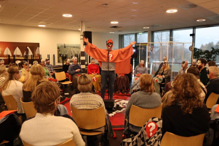 En mann kledd i en rød kappe og sort og rød maske foran øynene, står i midten. Rundt han sitter mennesker kledd i masker. Foto: Marthe Haugdal.