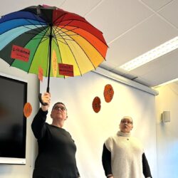 To damer i et klasserom. Den ene holder en fargerik paraply.