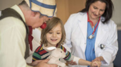 En sykehusklovn viser frem en bamse til et barn. I bakgrunnen ser vi en lege. Foto: https://www.rednoses.org/