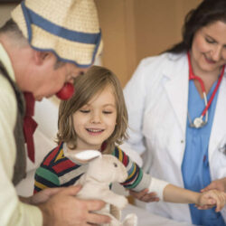 En sykehusklovn viser frem en bamse til et barn. I bakgrunnen ser vi en lege. Foto: https://www.rednoses.org/