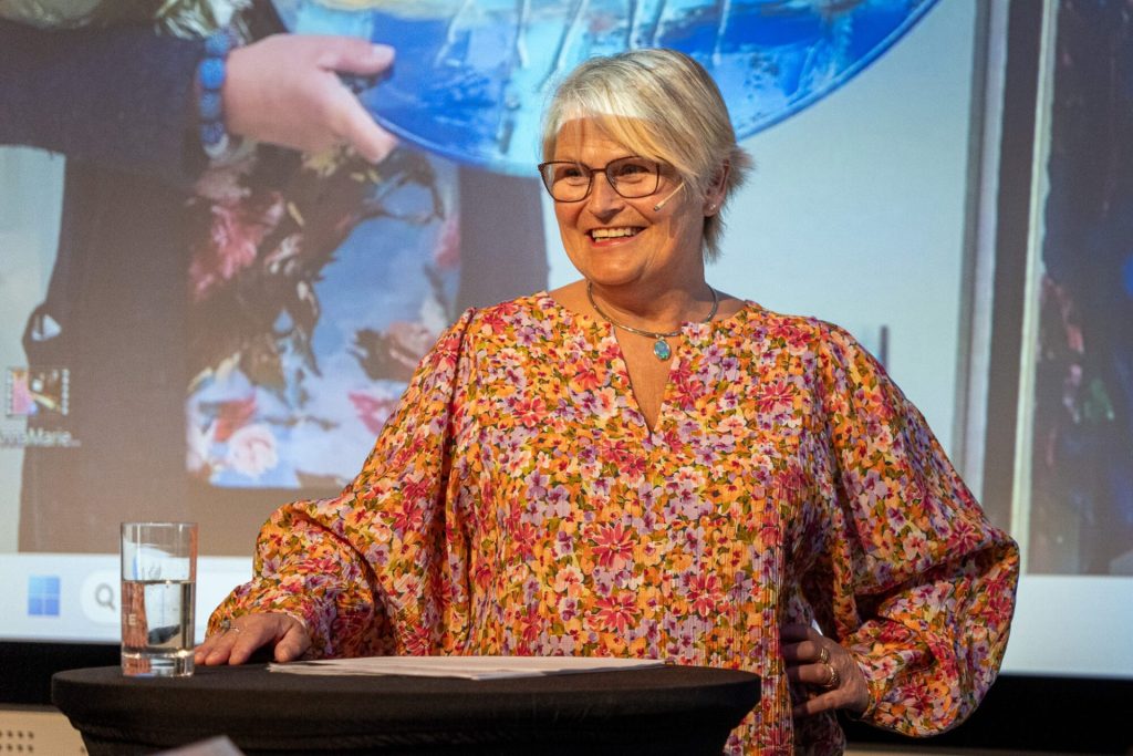 Anna Marie Laugsand står ved et talerbord. Hun har lyst hår, briller og en blomstrete bluse. Hun smiler. Foto: Alexandra Dahlen.