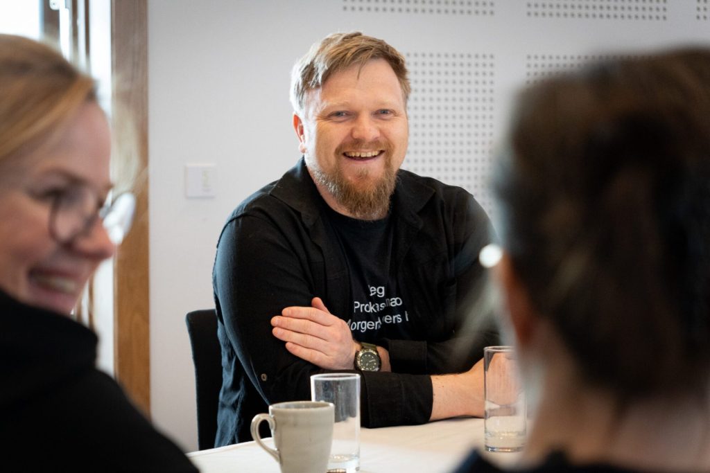 En mann sitter og smiler ved et bord. I forgrunn kan vi skimte to andre personer. Foto: Alexandra Dahlen.