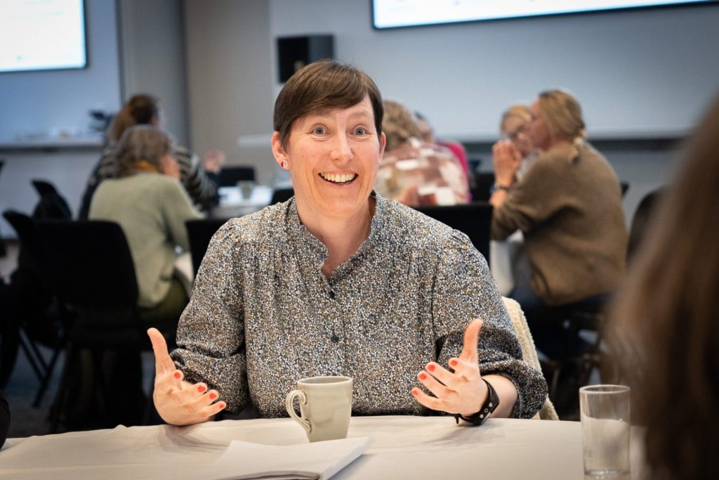 Mariann Lødemel sitter ved et bord. Foran henne står en kaffekopp. Hun smiler. Hun har kort, mørkt hår, en går bluse. I bakgrunn ser vi andre personer som sitter samlet. Foto: Alexandra Dahlen.