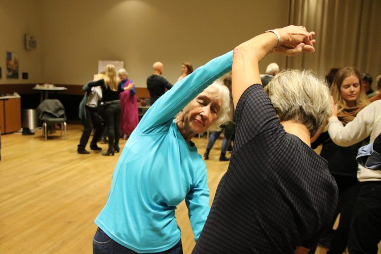 To kvinner danser. Den ene kvinnen har lukkede øyne, hvitt, kort hår, og en lysblå genser. Den andre kvinnen ser vi bakfra. Hun har sort t-skjorte, og grått, kort hår. Bak kvinnen ser vi flere mennesker som danser. Foto: Marthe Haugdal