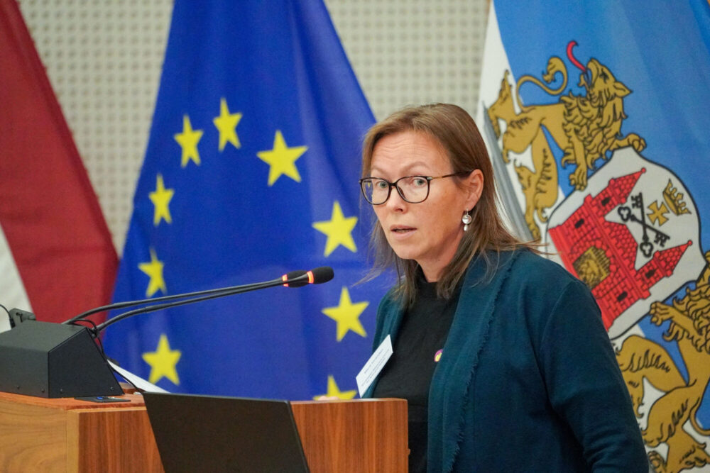 Marit Stranden står ved en talerstol. Marit har mørkeblondt, skulderlangt hår, briller og har på seg en mørk blazer. I bakgrunnen ser vi ulike flagg.