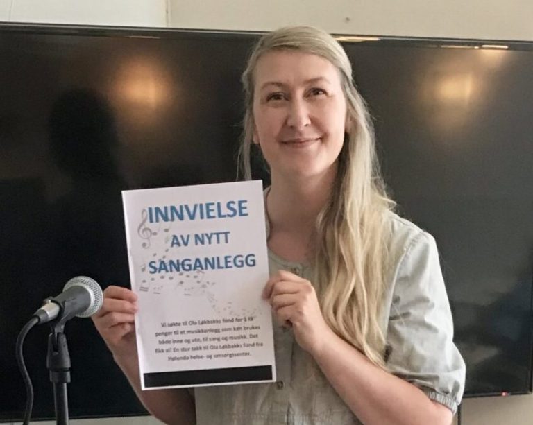 Charlotte Stav smiler til kamera. Hun har langt, lyst hår og holder en plakat i hendene. Ved siden av henne står en mikrofon.