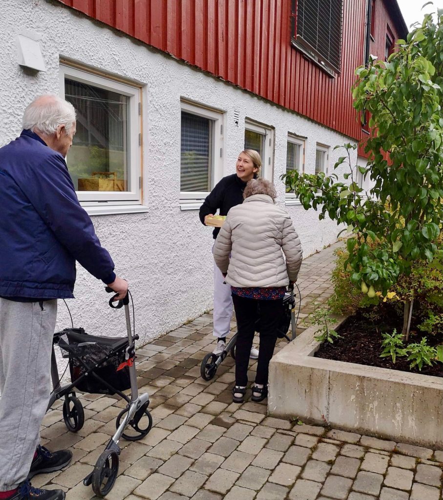 En ung kvinne er ute sammen med en eldre mann og en eldre kvinne. De går sammen langs et hus. De to eldre menneskene har rullator.