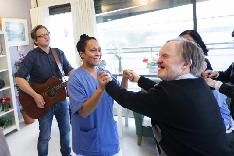 En helsearbeider kledd i blå skjorte danser med en mann. Bak dem ser vi flere som danser og en mann med gitar. Foto: Martin Lundsvoll.