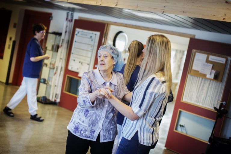 En eldre kvinne kledd i lilla bluse og med skaut på hodet, danser med en yngre kvinne med langt, blondt hår. I bakgrunn ser vi flere helsearbeidere. Foto: Martin Lundsvoll.