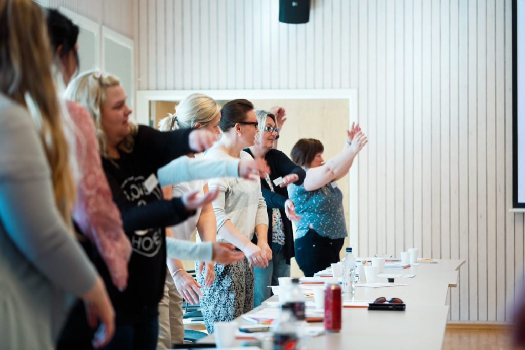 Flere kvinner står i et lyst rom foran pulter som er satt ved siden av hverandre. De rører på hendene sine.