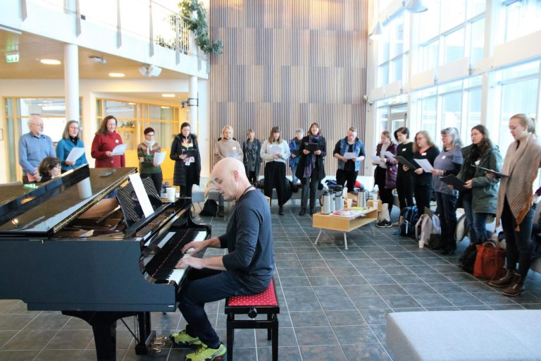 Anders Dalane spiller piano. I bakgrunnen ser vi flere mennesker som står ved siden av hverandre og synger.