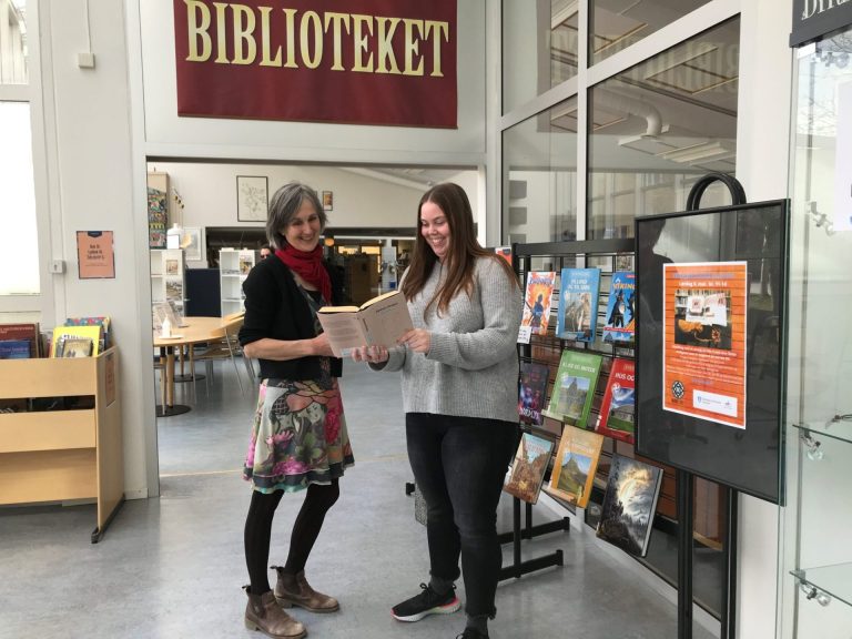 To kvinner står i et bibliotek ved siden av hverandre og smiler. Kvinnen til høyre holder en bok som de begge ser i.