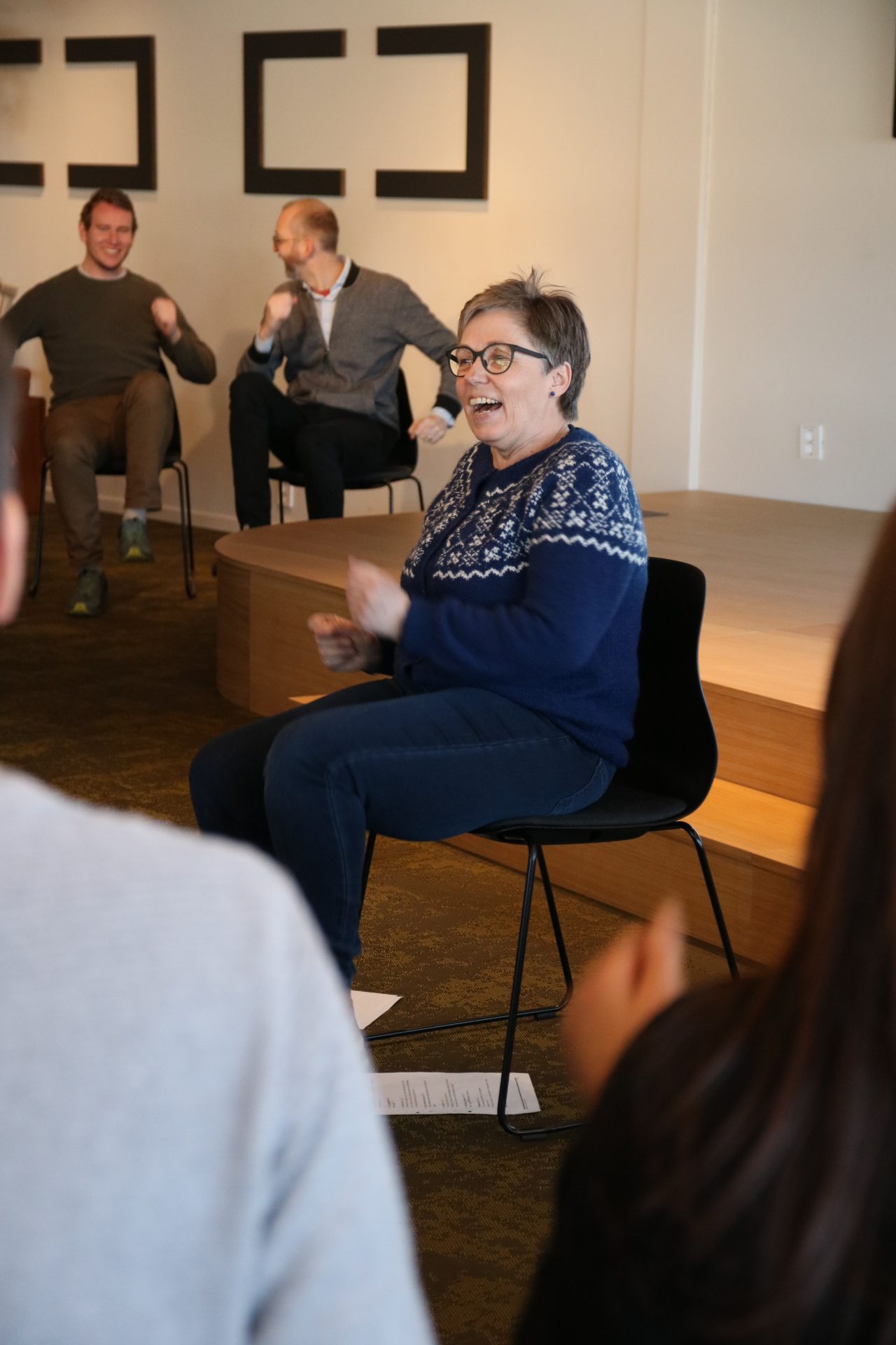 En kvinne med kort hår og briller, sitter på en stol. Hun smiler. Hun har på seg en blå strikket genser med hvitt mønster. I bakgrunnen ser vi flere personer som sitter på stoler og smiler. Foto: Alexandra Dahlen.