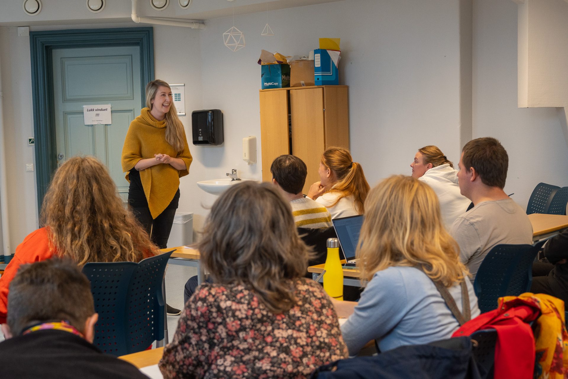 Charlotte Stav har langt, lyst hår og er kledd i en okergul poncho og svart bukse. Hun står foran en klasse i et klasserom. Foto: Alexandra Dahlen.