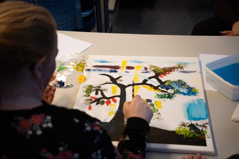 En kvinne holder et sugerør i høyre hånd og en malepalett i venstre. Foran henne ligger et maleri hun maler på. Foto: Alexandra Dahlen.