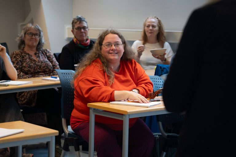 Kvinne med langt krøllet hår, briller og oransje genser sitter ved pulten i et klasserom. Foto: Alexandra Dahlen