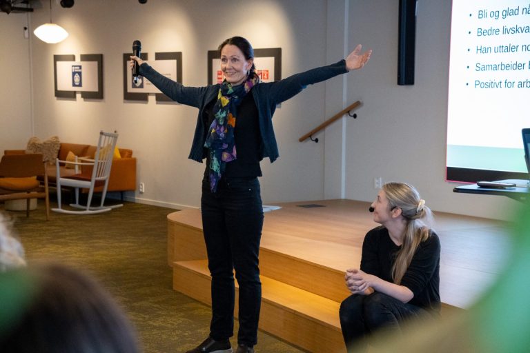 Katrine Homdrum står med armene rett ut til siden. Hun har mørkt hør i en strikk. Har på seg mørk bukse, mark cardigan og en fargerik topp. Ved siden av henne sitter Charlotte Stav. Foto: Alexandra Dahlen.