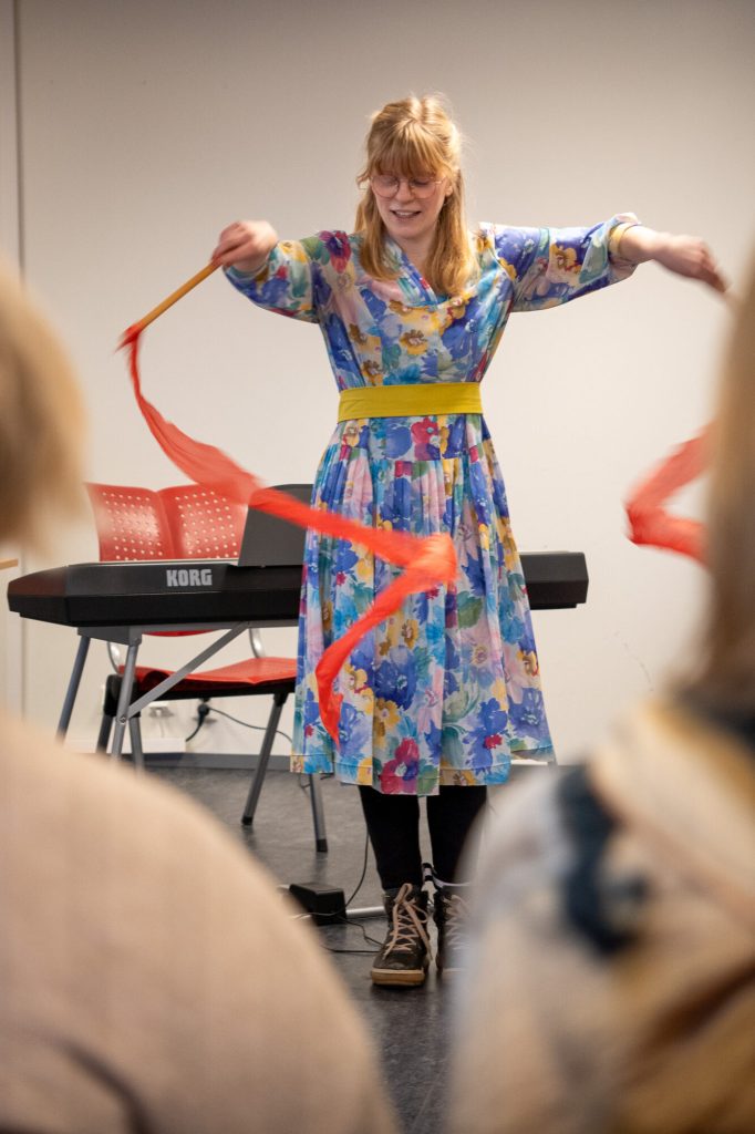 Kvinne står med en pinne i hver hånd som har et rødt silkebånd knyttet fast i seg. hun beveger pinnene slik at silketråden lager en spiral form. Foto: Alexandra Dahlen.