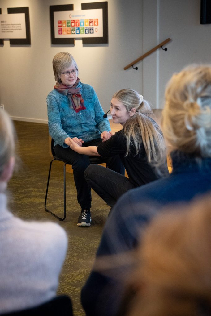 To damer står foran en forsamling. Den ene sitter på en stol, den andre sitter på huk foran. Foto: Alexandra Dahlen