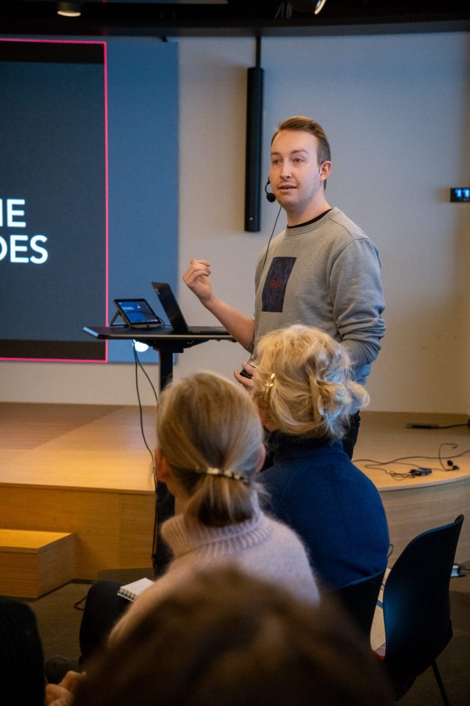 En mann med kort hår og lys genser står foran flere mennesker. Han har en mygg (mikrofon) på seg. Foto: Alexandra Dahlen.