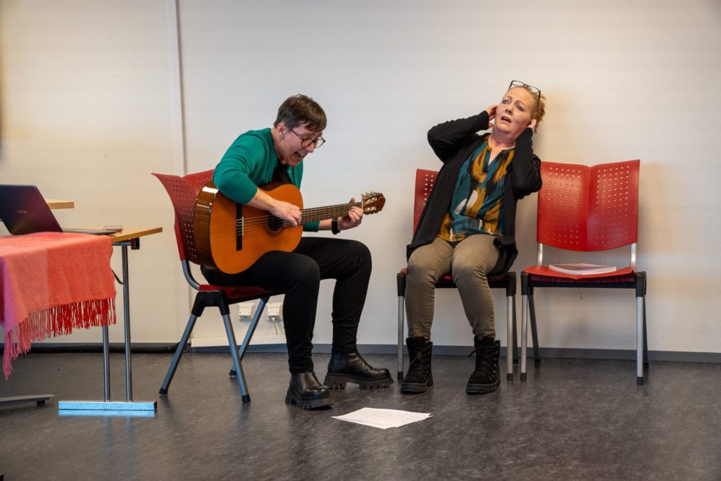 To kvinner sitter på hver sin stol. Dene ene kvinnen spiller gitar og synger. Den andre kvinnen holder seg for ørene. Foto: Alexandra Dahlen