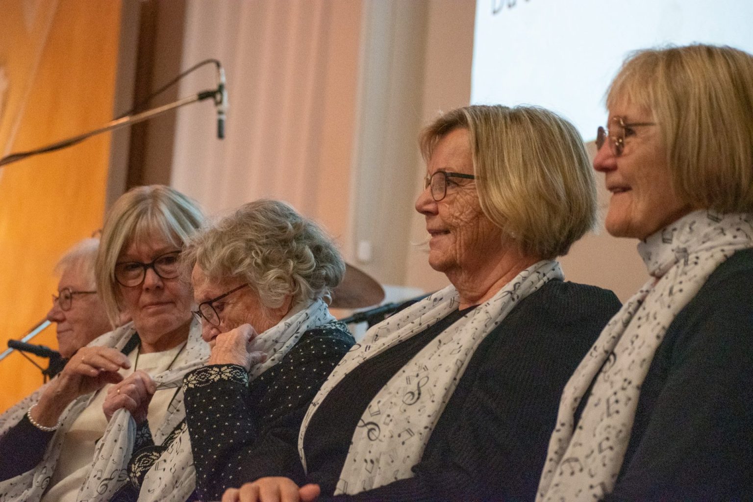 Fem kvinner sitter samlet. De har alle lyst hår og har på seg et hvitt skjerf med sorte noter på. Foto: Gonzalo Sentana.