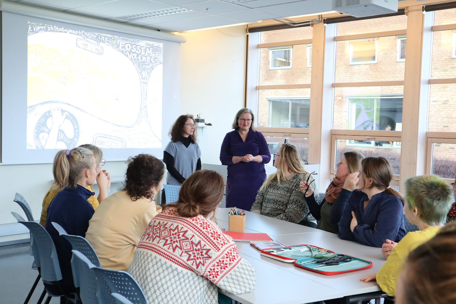 Studenter ved videreutdanningen "Kunst og kultur i helse og omsorg" sitter sammen ved et bord. På bordet ligger ark, saks og blyanter i ulike farger. To kvinner står og snakker foran de andre. Foto: Marit Stranden.