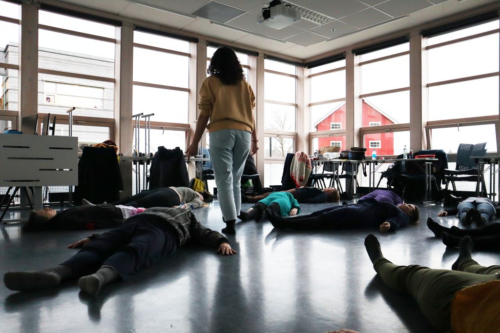 En kvinne går rundt på gulvet i et klasserom. Rundt henne ligger mange personer på gulvet. Foto: Marit Stranden.