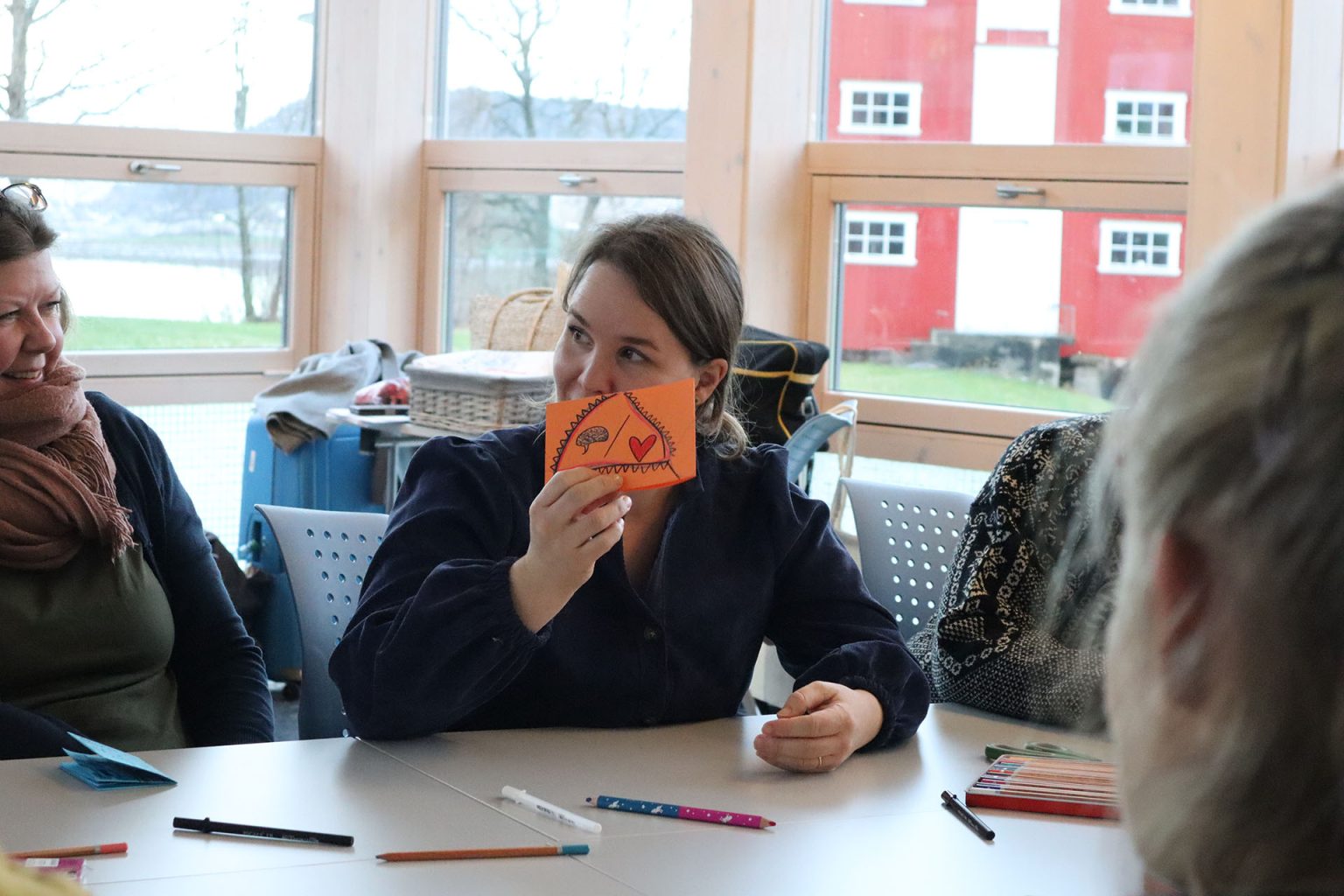 En kvinne sitter sammen med andre ved et bord. På bordet ligger det blyanter i ulike farger. Kvinnen har mørkt hår i en strikk og mørkeblå skjorte. Hun holder et oransje papir som det er tegnet på foran ansiktet. Foto: Marit Stranden.