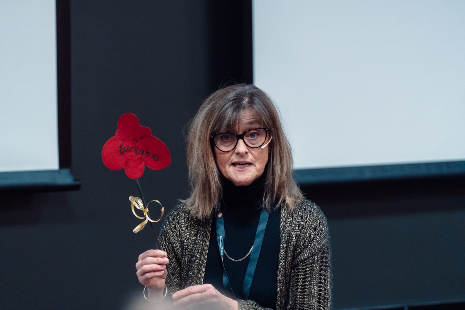 Ragnhild Skille har skulderlangt hår, briller og en sort genser på seg med en cardigan over. Hun holder i en papirblomst. Foto: Christian Trustrup, TA-Kreativ.