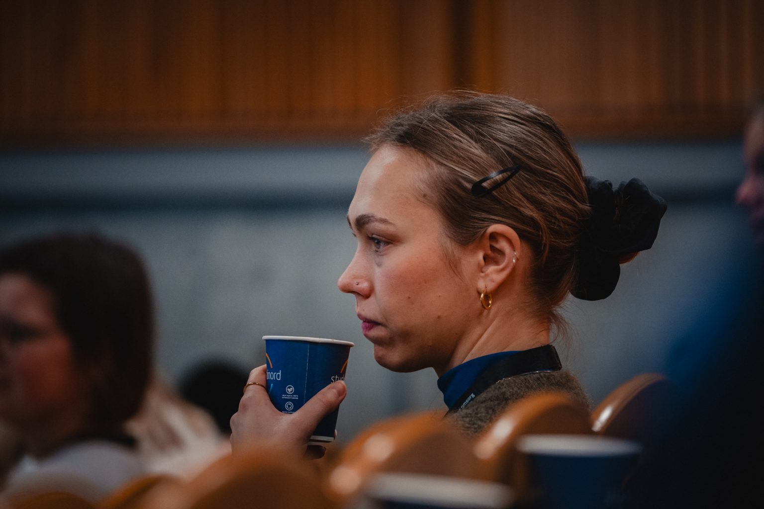 En kvinne sitter sammen med andre i en forelesningssal. Kvinnen har oppsatt hår og holder en kaffekopp. Foto: Christian Trustrup, TA-Kreativ.