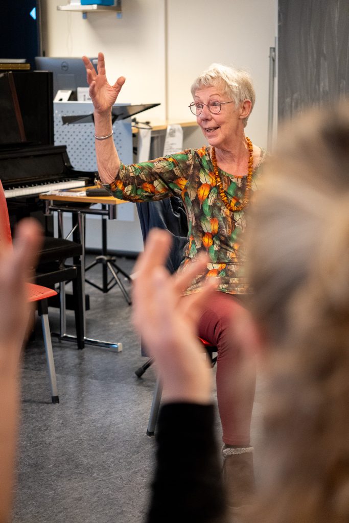 En eldre kvinne sitter på en stol. Hun har kort, lyst hår og briller. Hun smiler og vinker med den ene armen. Foto: Alexandra Dahlen.