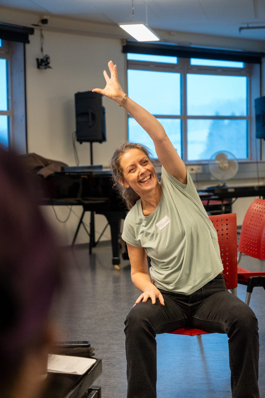 En kvinne med hestehale, grønn t-skjorte og sort bukse, sitter på en stol. Hun smiler og strekker venstre hånd i luften mens hun lener seg til siden. Foto: Alexandra Dahlen.