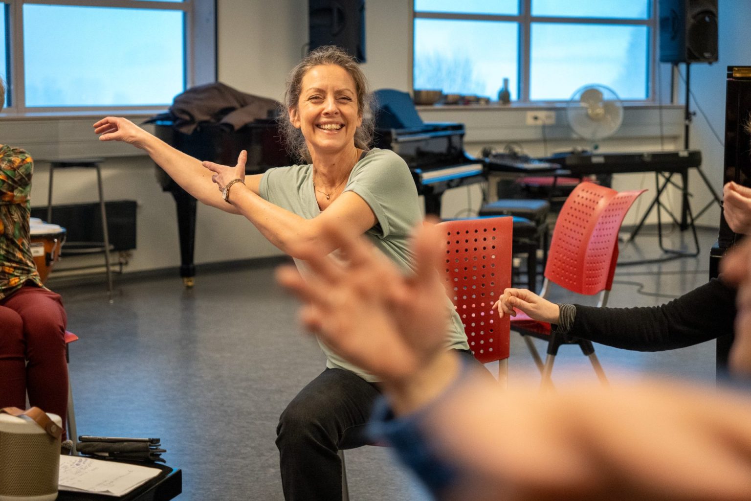 En kvinne med hestehale, grønn t-skjorte og sort bukse, sitter på en stol. Hun smiler mens hun slenger armene til høyre side. Foto: Alexandra Dahlen.