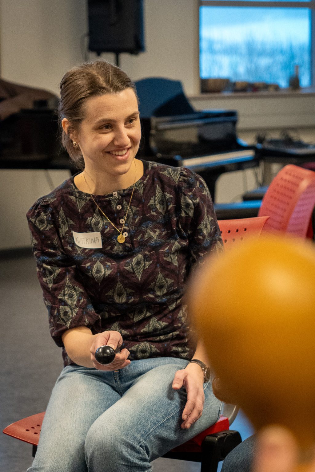 En kvinne sitter på en stol. Hun har hestehale, mønstret bluse og dongeribukse. Hun holder et rytmeinstrument i hånden. Foto: Alexandra Dahlen.