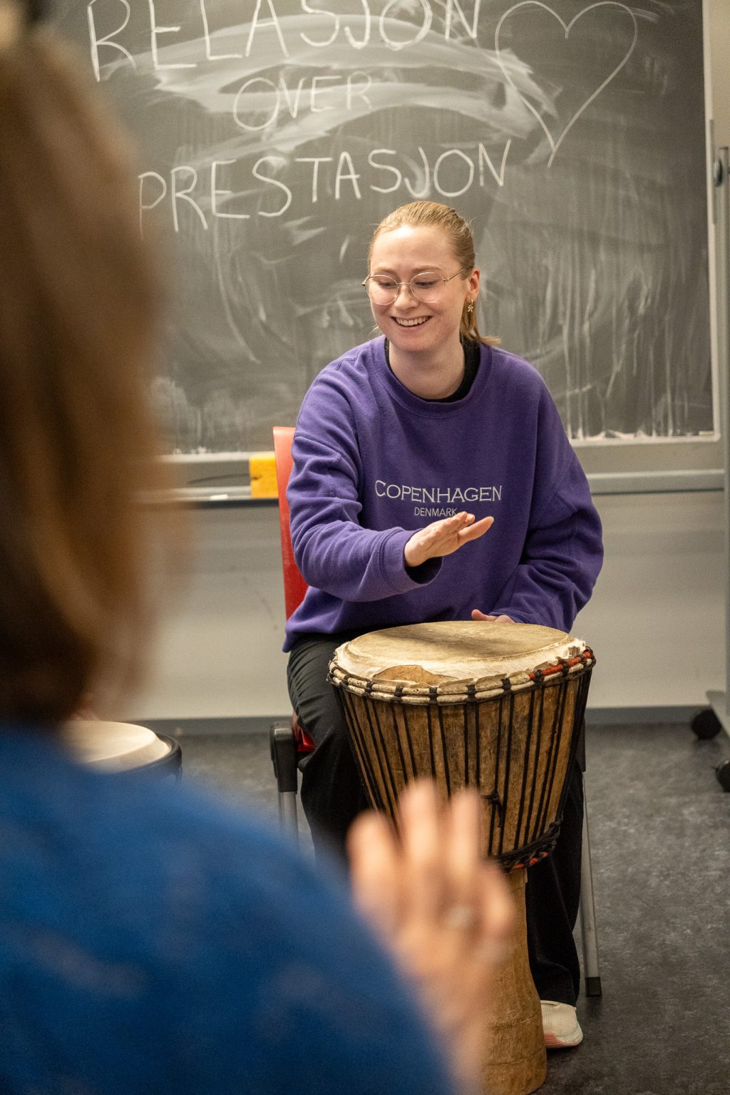 En kvinne sitter på en stol og slår på en tromme. Hun har lyst hår i en hestehale, briller, lilla genser og sort bukse. Hun smiler. Foto: Alexandra Dahlen.