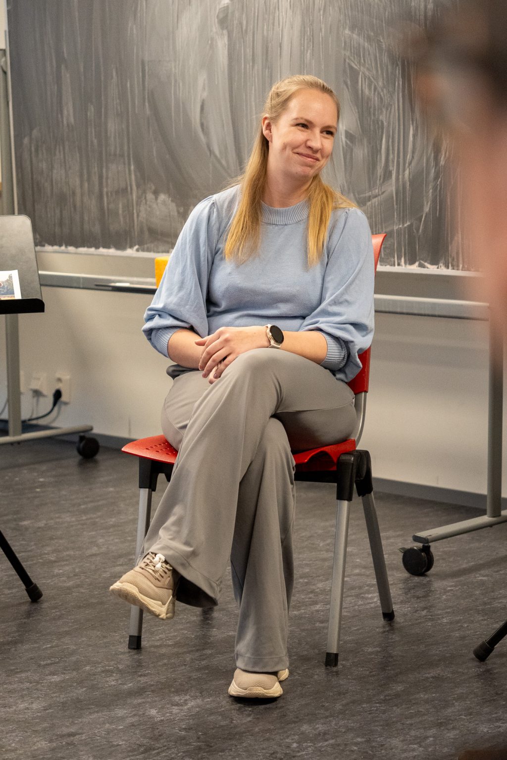 En kvinne med langt, lyst hår, blå genser og grå bukse, sitter på en stol og smiler. Foto: Alexandra Dahlen.