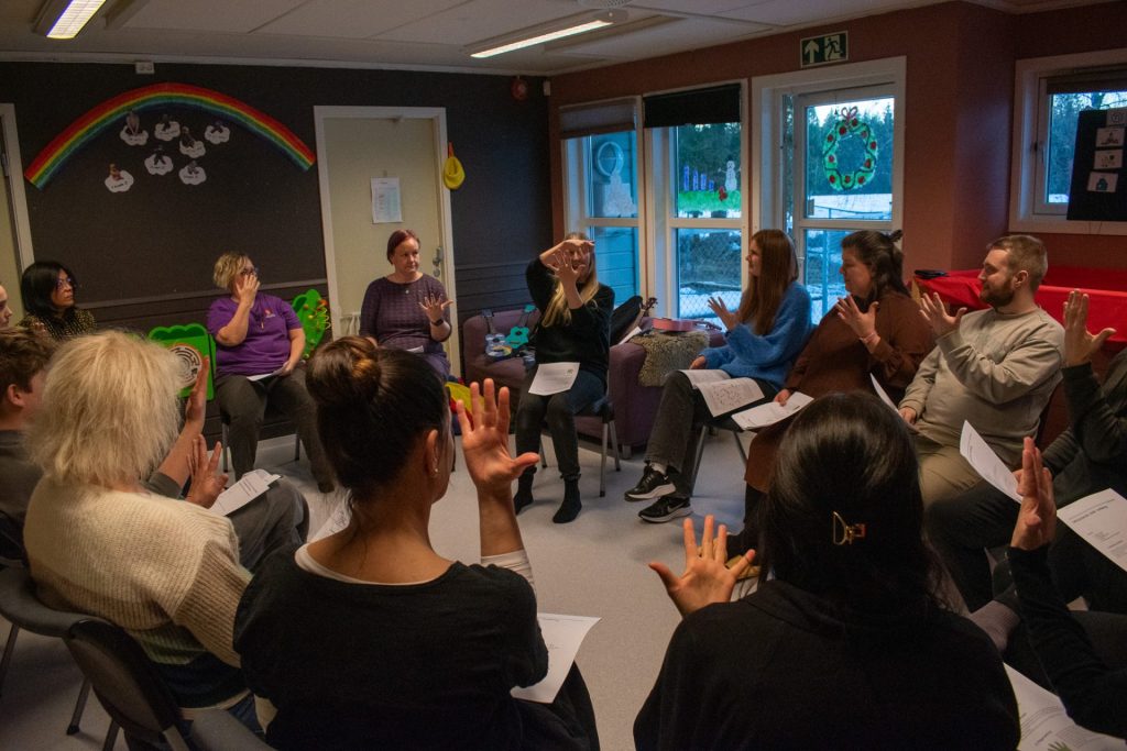 En gruppe voksne sitter på stoler som er plassert i en sirkel. Alle holder en hånd foran seg og ser på Charlotte Stav som instruerer. Foto: Gonzalo Sentana.