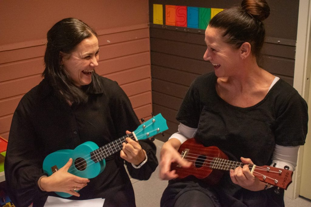 To kvinner med mørkt hår sitter ved siden av hverandre. De ser på hverandre og ler. De holder begge i hver sin ukulele. Foto: Gonzalo Sentana.