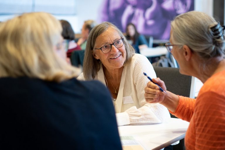 Hennie Skaufel sitter og smiler til Kristin Nøst som sitter rett over henne. Foto.