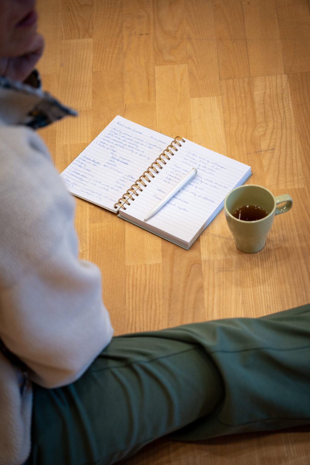 En person sitter på gulvet. Foran ligger en notatbok og en kaffekopp. Foto.