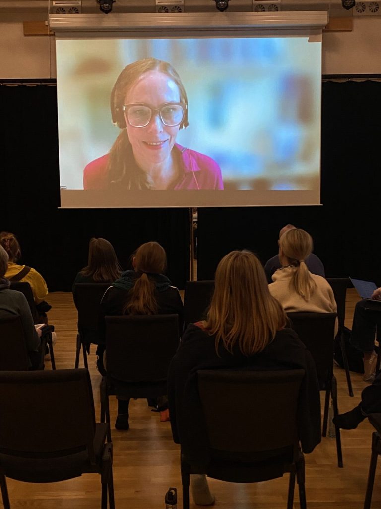 På bildet ser vi flere personer sitter på stoler i en sal. De ser på en skjerm og på skjermen ser vi Anita Jensen. Bildet er tatt under danseseminaret "Å skape meningsfulle dager". Foto: Alexandra Dahlen.