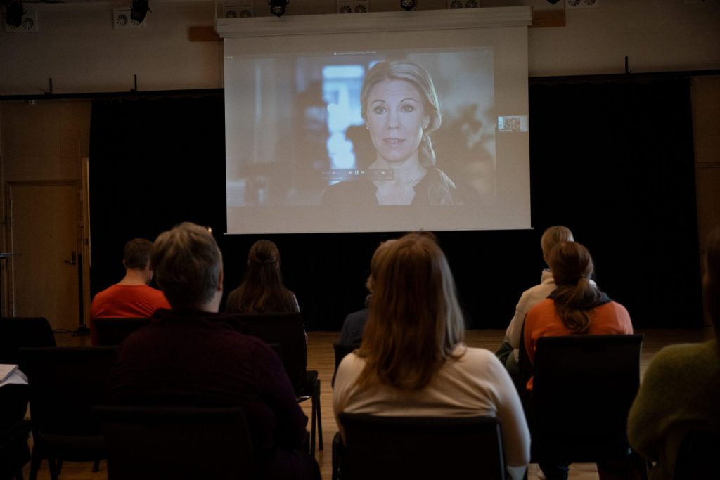 På bildet ser vi flere personer sitter på stoler i en sal. De ser på en skjerm og på skjermen ser vi Anna Duberg. Bildet er tatt under danseseminaret "Å skape meningsfulle dager". Foto: Alexandra Dahlen.