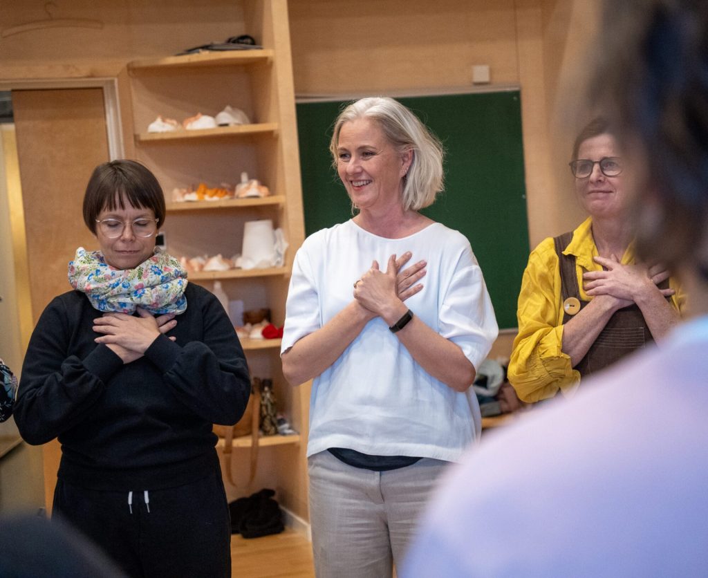 Elli Isokoski og Kari Anne Vadstensvik Bjerkestrand står ved siden av hverandre og holder hendene på brystet. Kari Anne smiler og Elli holder øynene lukket. Dlere andre kvinner står ved siden av dem. Bildet er tatt under danseseminaret "Å skape meningsfulle dager". Foto: Alexandra Dahlen.