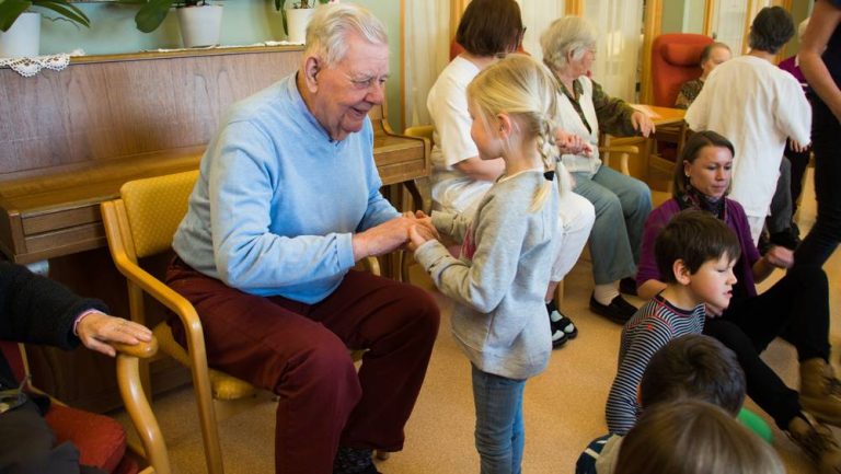 En eldre mann sitter på en stol. Han holder hendene til ei lita jente. Foto.