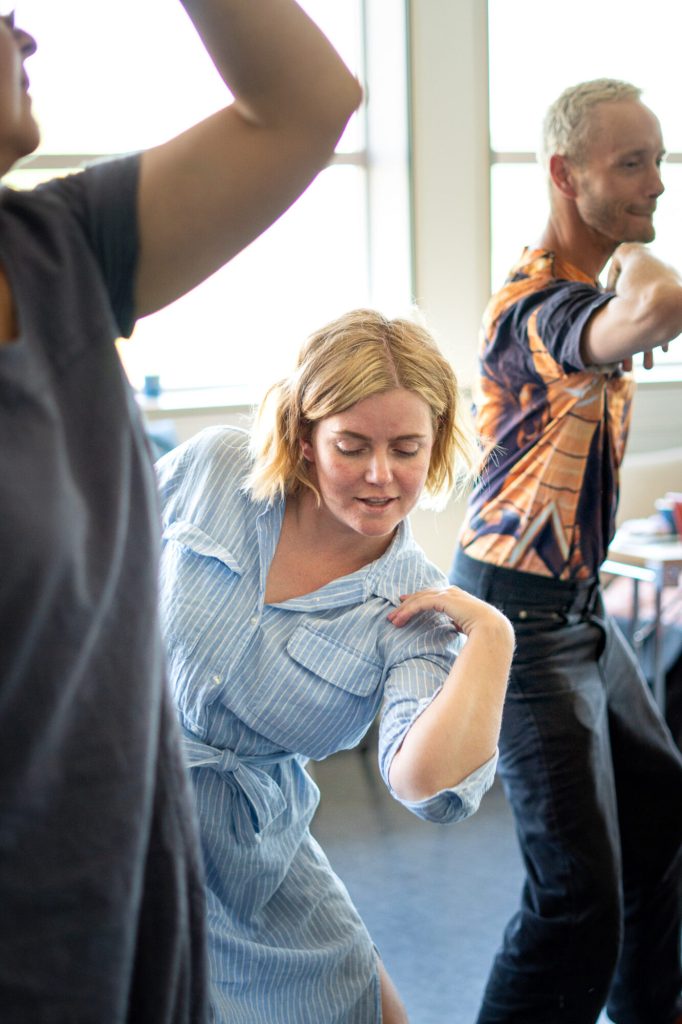 Guro Gulstad danser. Hun har lyst, kort hår og en lyseblå skjortekjole. I bakgrunn og i forgrunn ser vi andre som danser. Foto: Alexandra Dahlen.