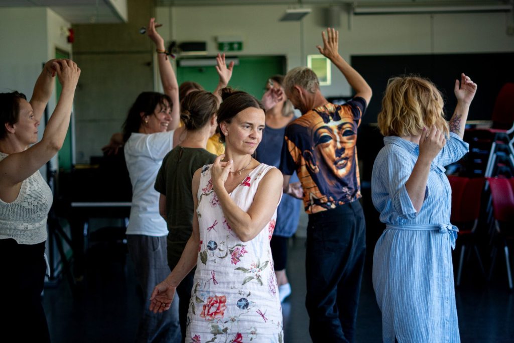 En gruppe mennesker danser sammen. I forgrunn kan vi sen en kvinne i lys kjole med blomster, mørkt hår som er oppsatt. Foto: Alexandra Dahlen.