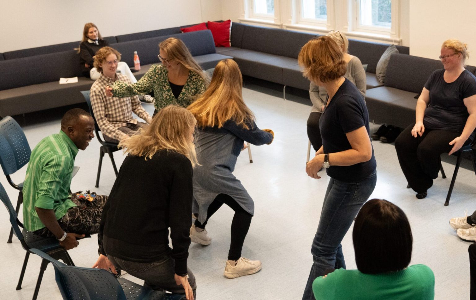 Flere personer sitter på stoler i en ring. Noen av dem løper rundt i ringen. Foto.