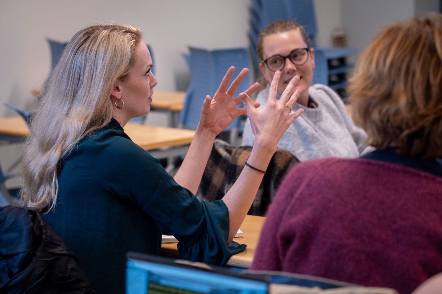 En kvinne sitter ved en pult i et klasserom og snakker. Foto.