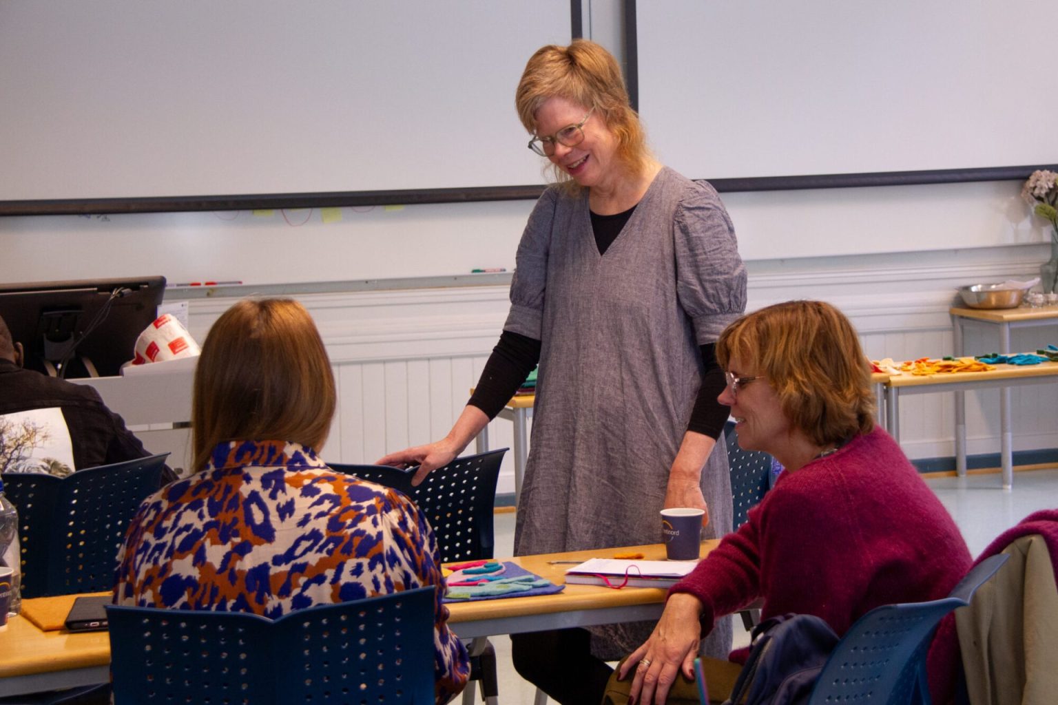 Anne Helga Henning står og snakker med to studenter som sitter ved pulten. Foto.